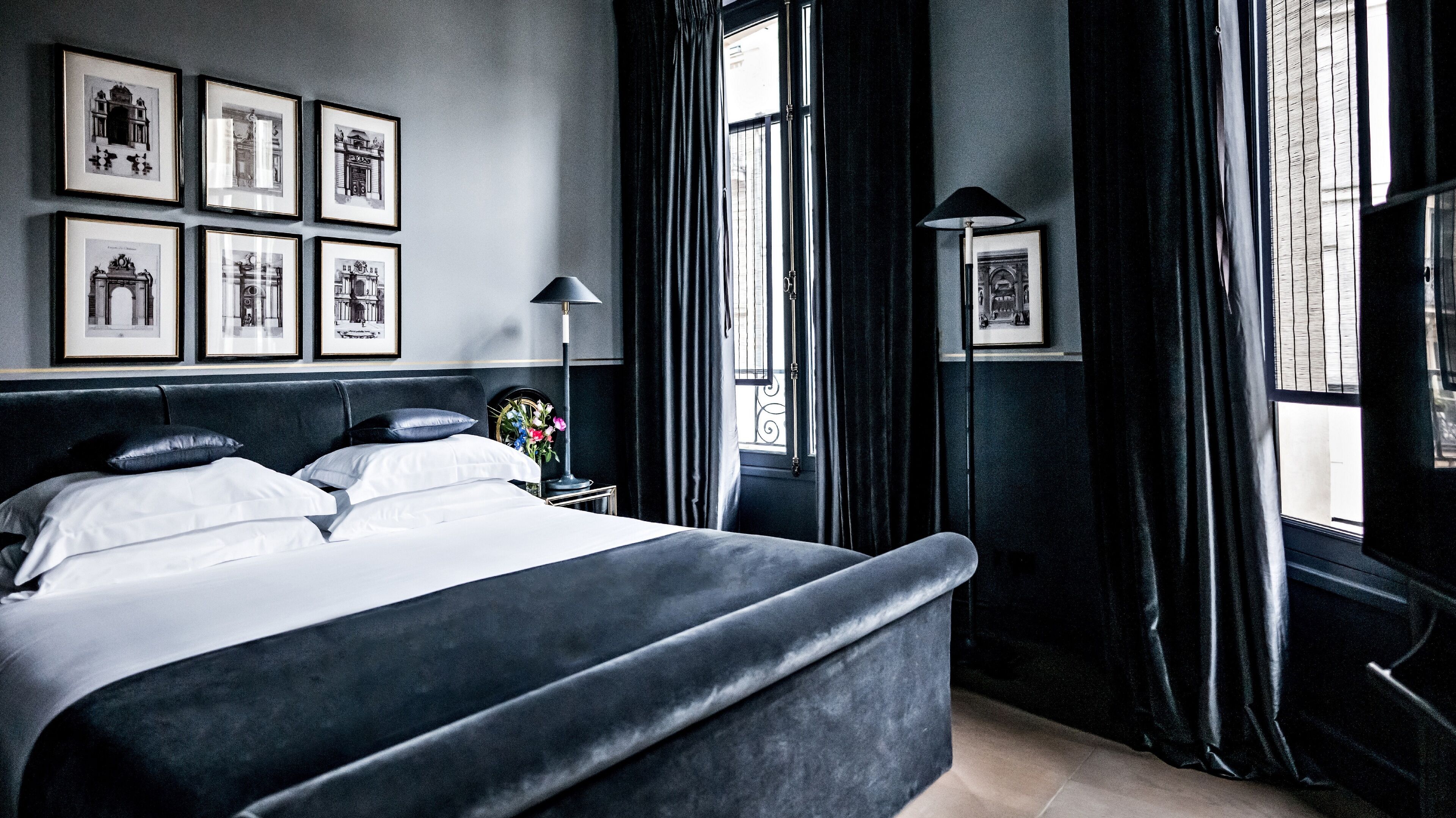 A neatly made bed with white linens and a dark headboard in a room with large windows, black curtains, and framed pictures on the wall.