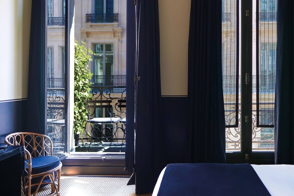 A hotel room with a bed, a desk, and a view of a balcony with outdoor furniture.