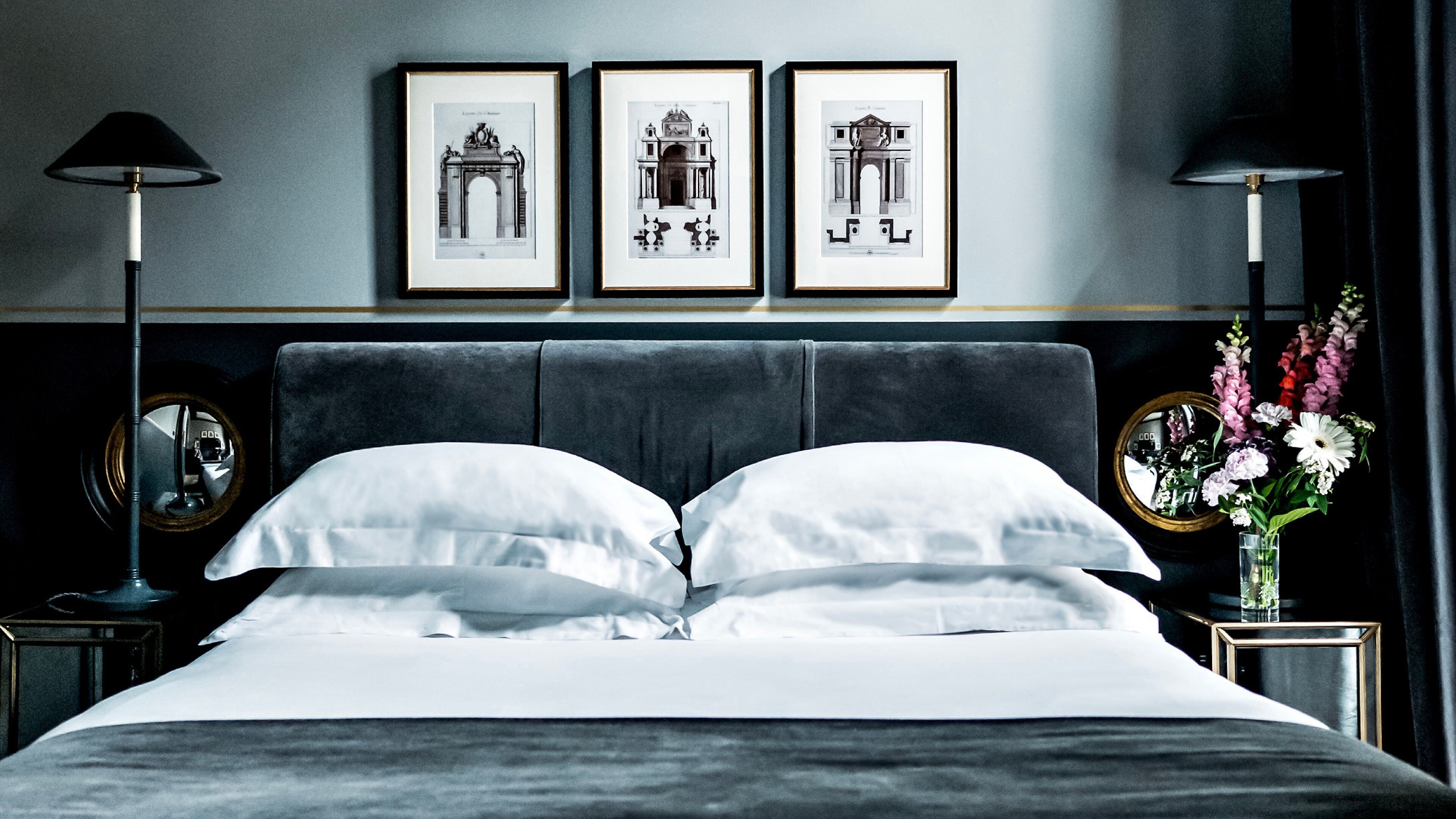 Una cama bien tendida con ropa blanca, un cabecero tapizado oscuro y tres dibujos arquitectónicos enmarcados en la pared de arriba.