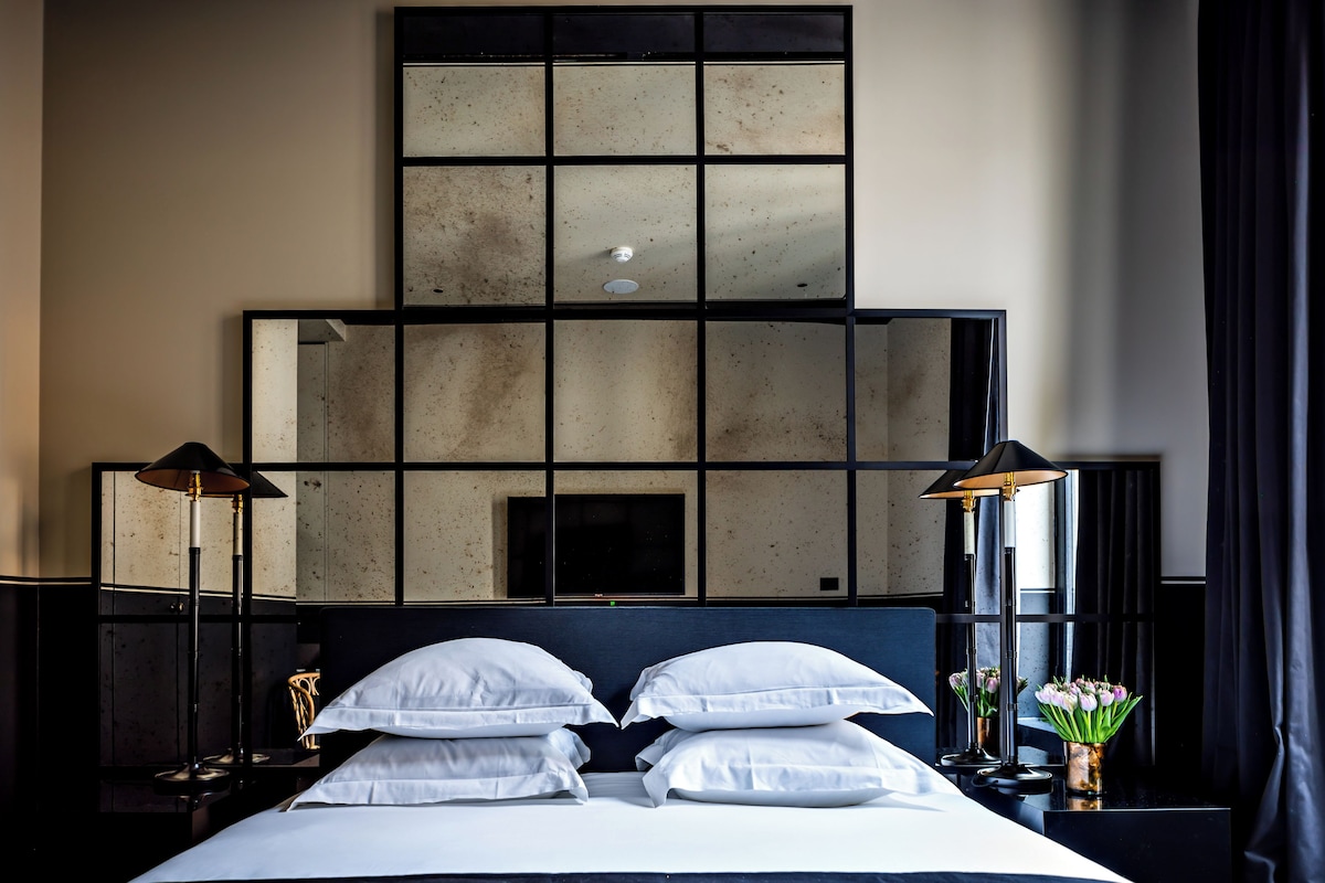 Een net opgemaakt bed met witte lakens, twee bedlampjes, een grote spiegel boven het bed en een bloevas op het nachtkastje.
