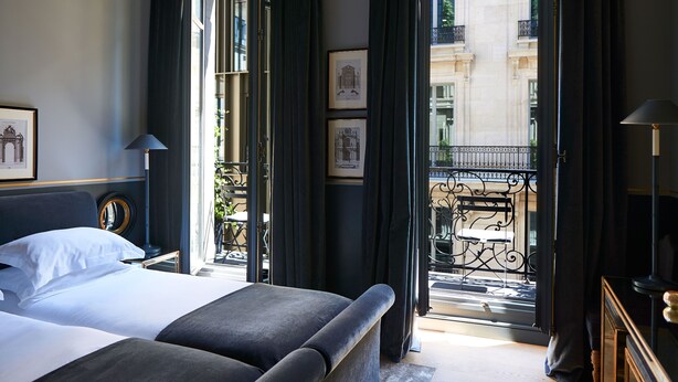 Habitación de hotel con una cama grande, una mesita de noche, un espejo y vistas a la ciudad a través de cortinas abiertas.
