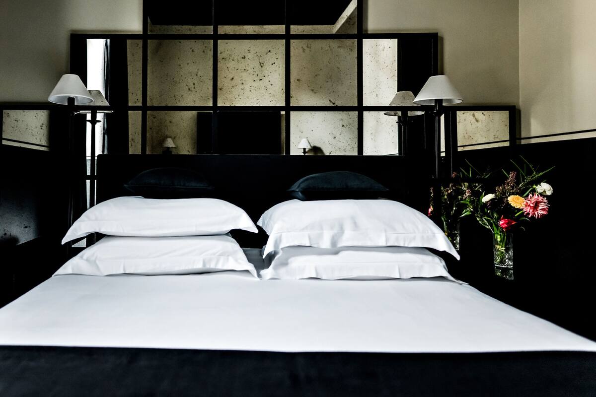 A hotel room with a large bed, two bedside lamps, a mirror above the bed, and a vase with flowers on the nightstand.