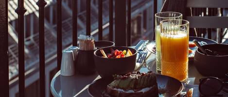 Una mesa de comedor con comida, un vaso de jugo de naranja y un bol de fruta en un balcón con vistas a edificios.