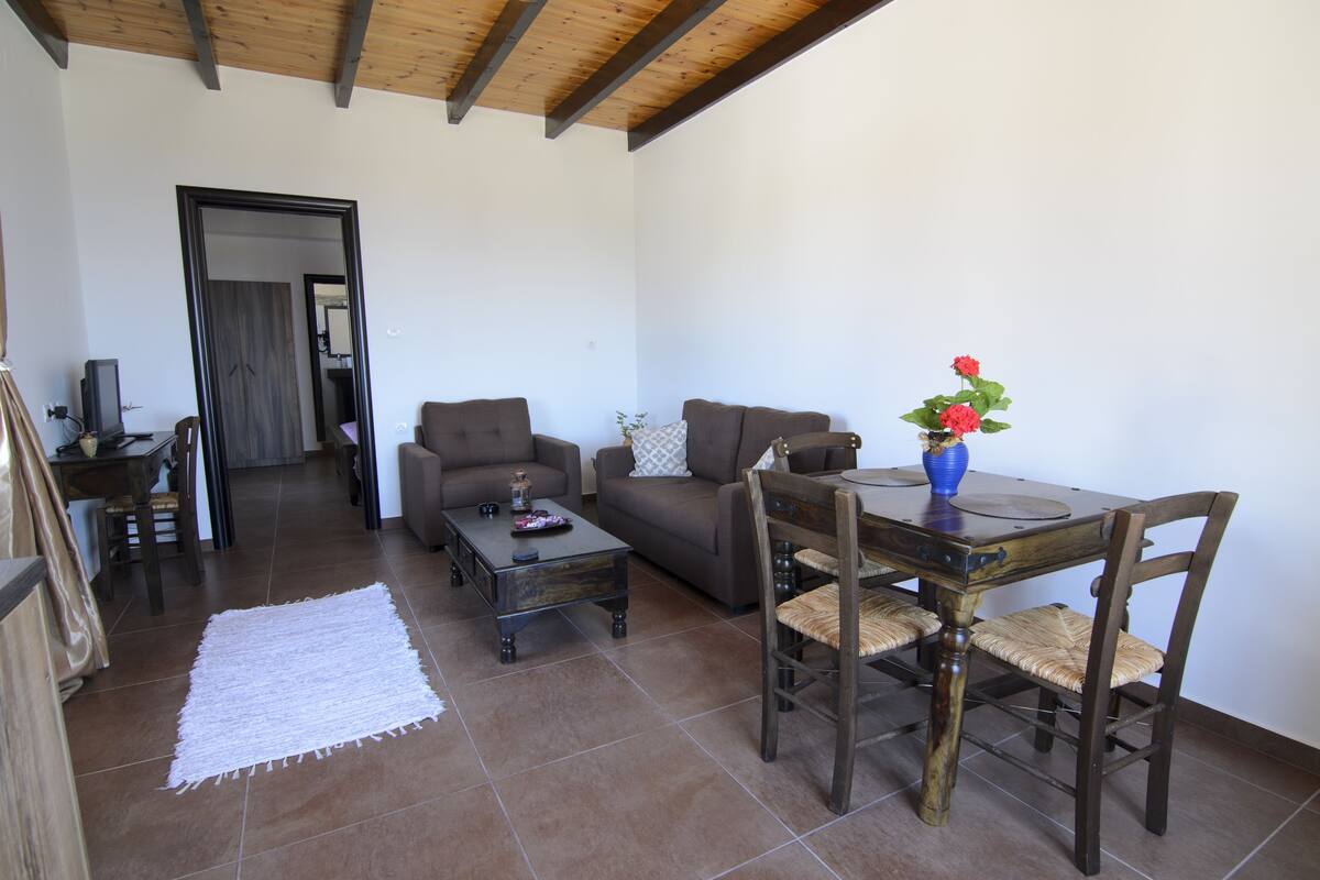 A living room with a sofa, coffee table, and dining table with chairs. There is a white rug on the floor and a door leading to another room.
