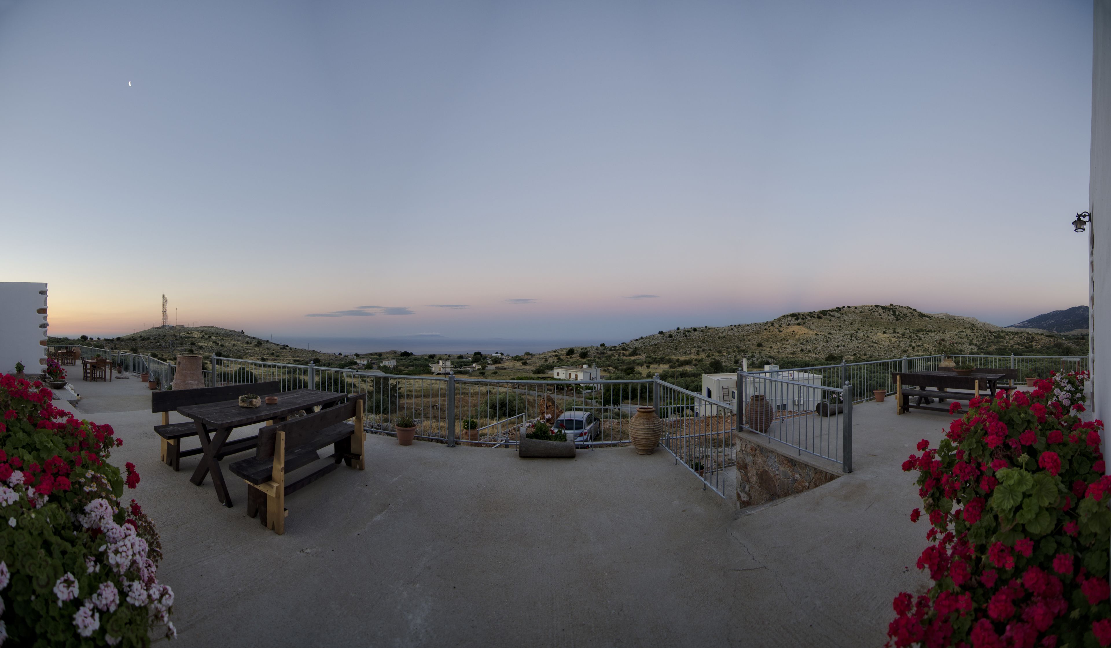 A terrace with a wooden table and benches, overlooking a hilly landscape with a distant view of the sea.