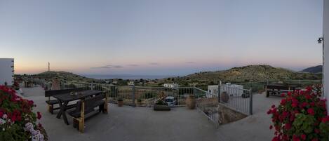 A terrace with a wooden table and benches, overlooking a hilly landscape with a distant view of the sea.