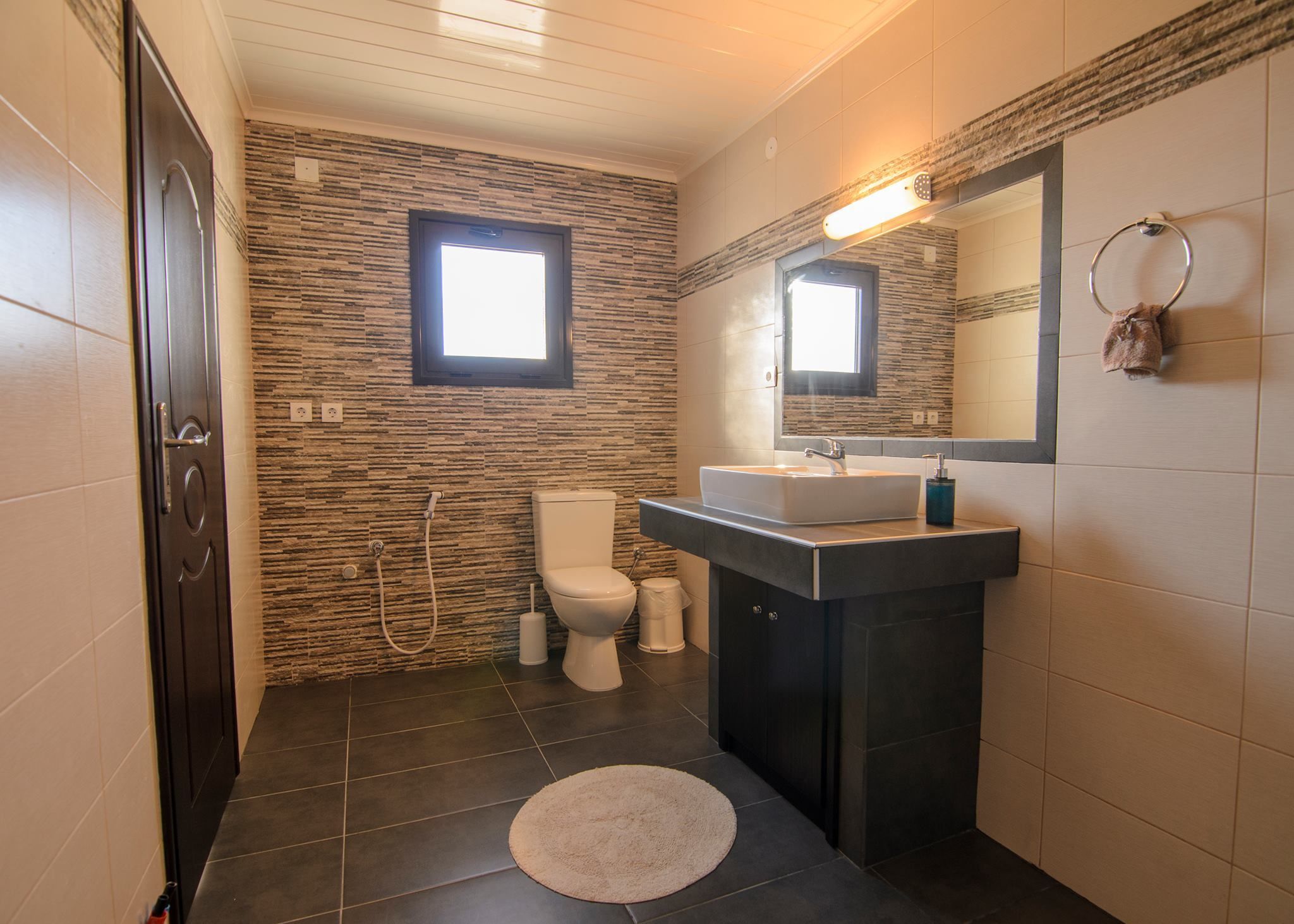 Une salle de bain au design moderne, avec un mur imitant la pierre, des toilettes blanches, un lavabo surmonté d’un miroir et une petite fenêtre.