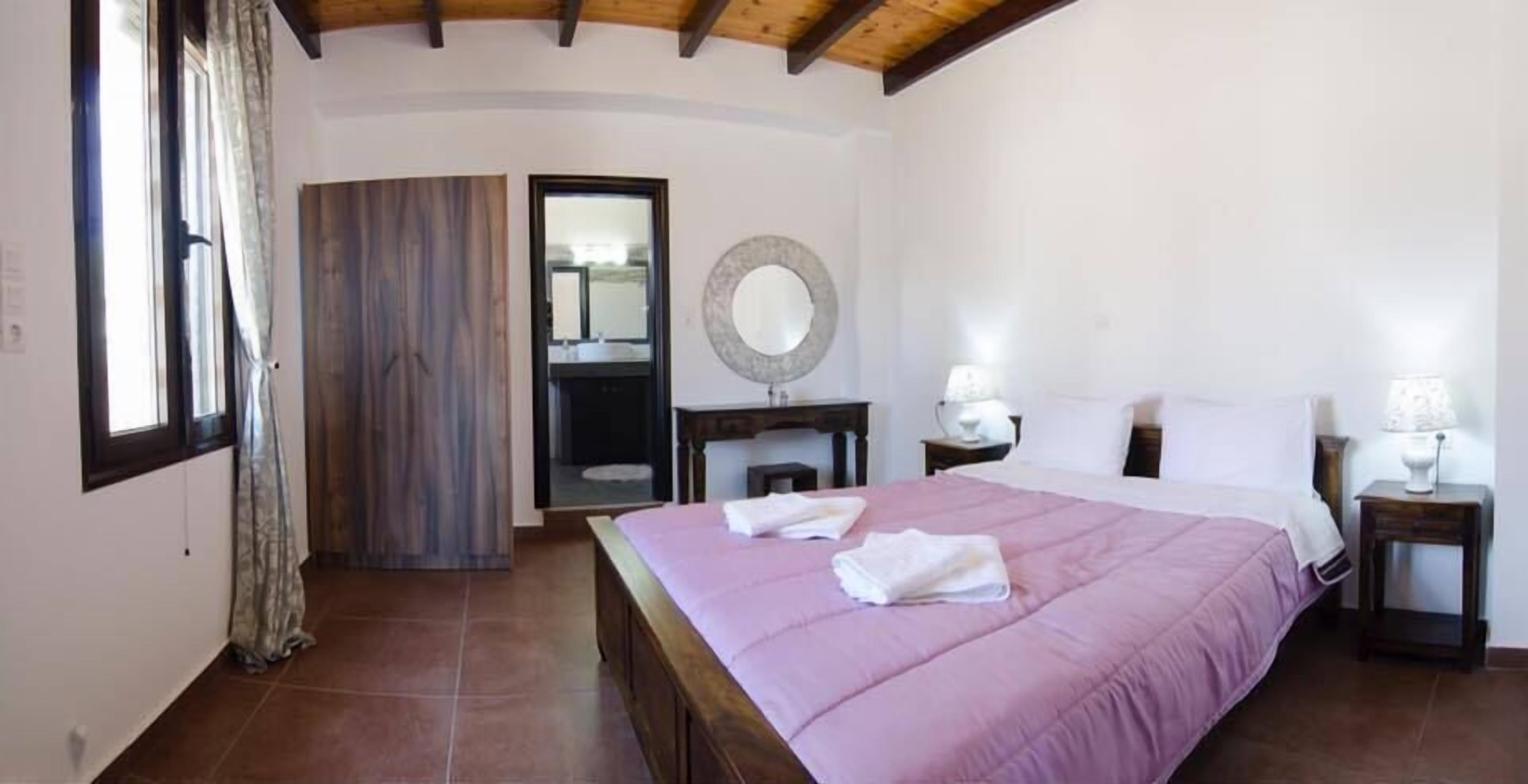 Un dormitorio con una cama grande, techo de madera, un espejo, una mesita de noche con lámpara y una ventana con cortinas.