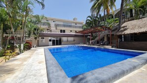 A swimming pool with a thatched roof structure, surrounded by palm trees and a building with multiple balconies. - Hotel DNA Cancun (Cancun)