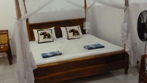 A bed with a wooden frame, white bedding, two pillows with animal designs, and a bedside table with a book. - Serene Home Stay (Udawalawa)