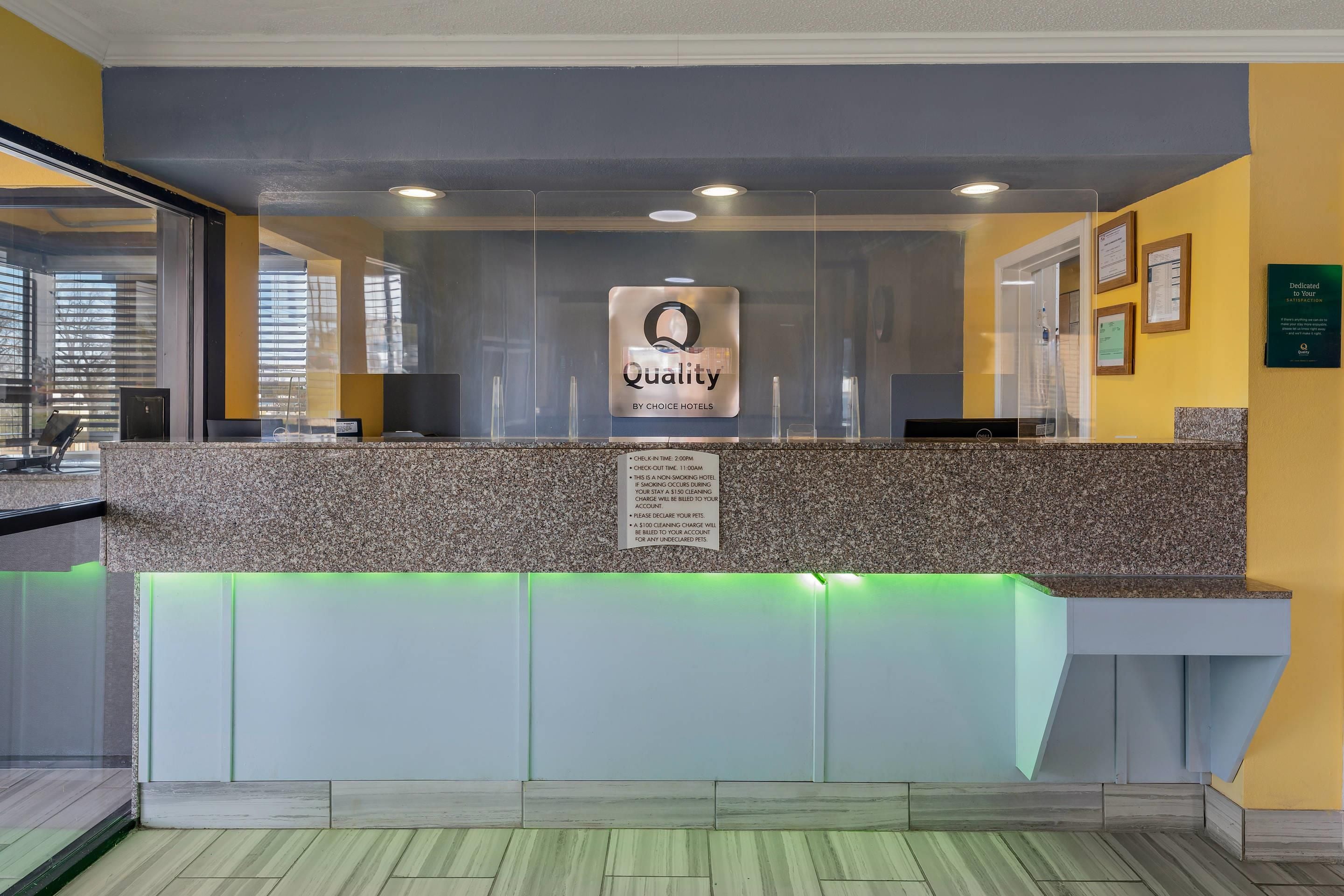 A hotel reception desk with a 'Quality by Choice Hotels' sign, a check-in/out time sign, and a no-smoking policy notice.