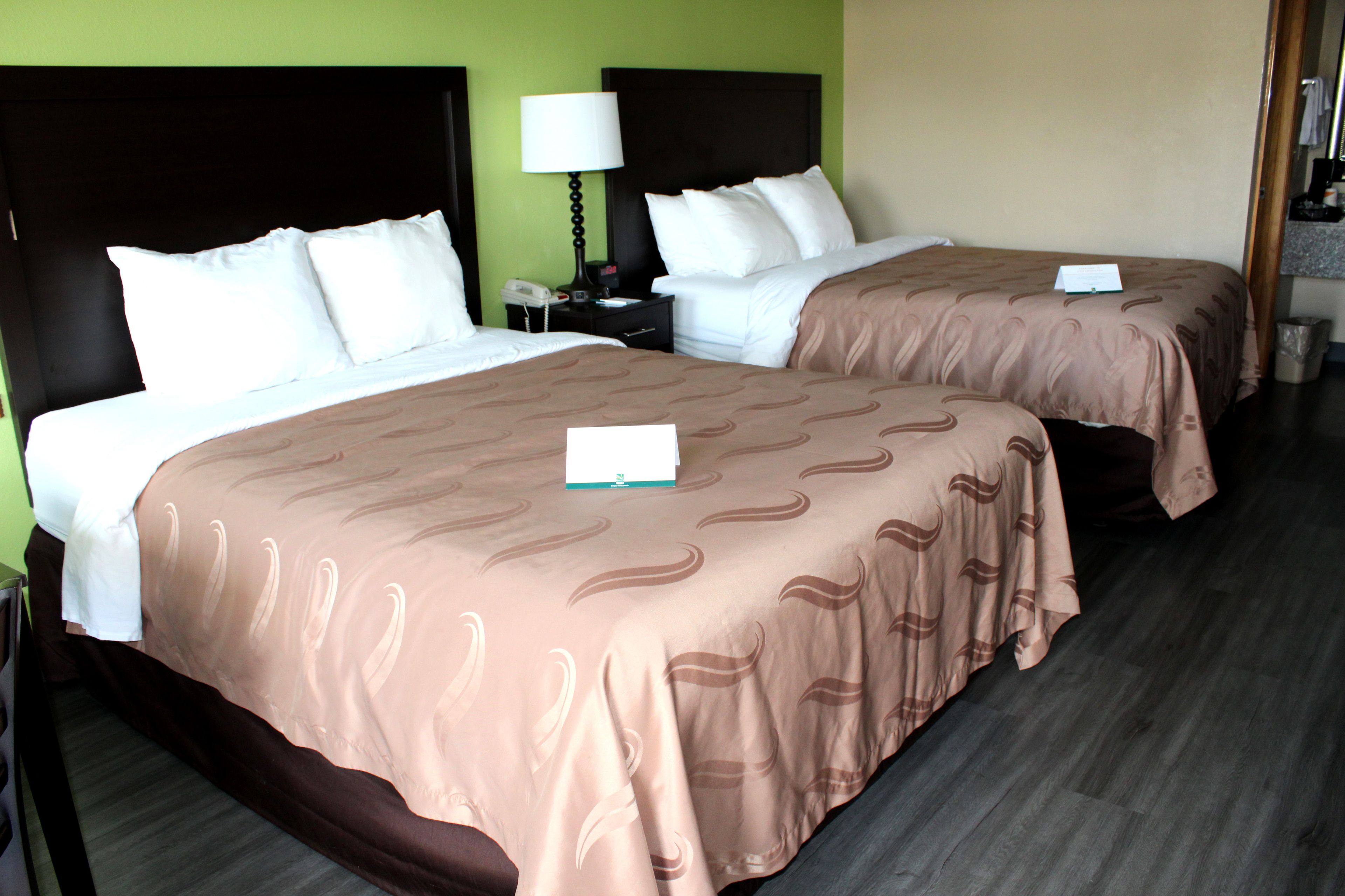 A hotel room with two beds, each with a brown bedspread and white pillows. There is a nightstand with a lamp and a phone between the beds. The room has a green wall and a wooden floor.
