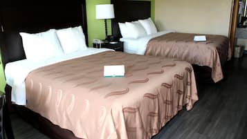 Quarto de hotel com duas camas, cada uma com colcha marrom e travesseiros brancos. Entre as camas, um criado-mudo com abajur e telefone. O quarto possui parede verde e piso de madeira.