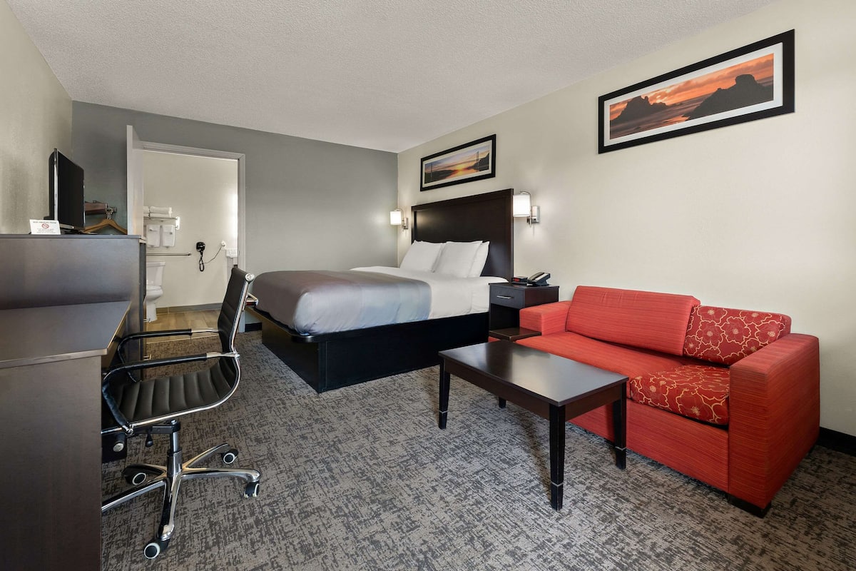 Habitación de hotel con cama, sofá rojo, escritorio con silla y un cuadro en la pared.