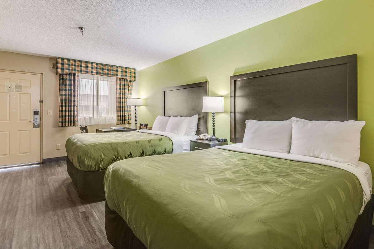 A hotel room with two beds, green bedding, a wooden headboard, a window with curtains, and a bedside table with a lamp.