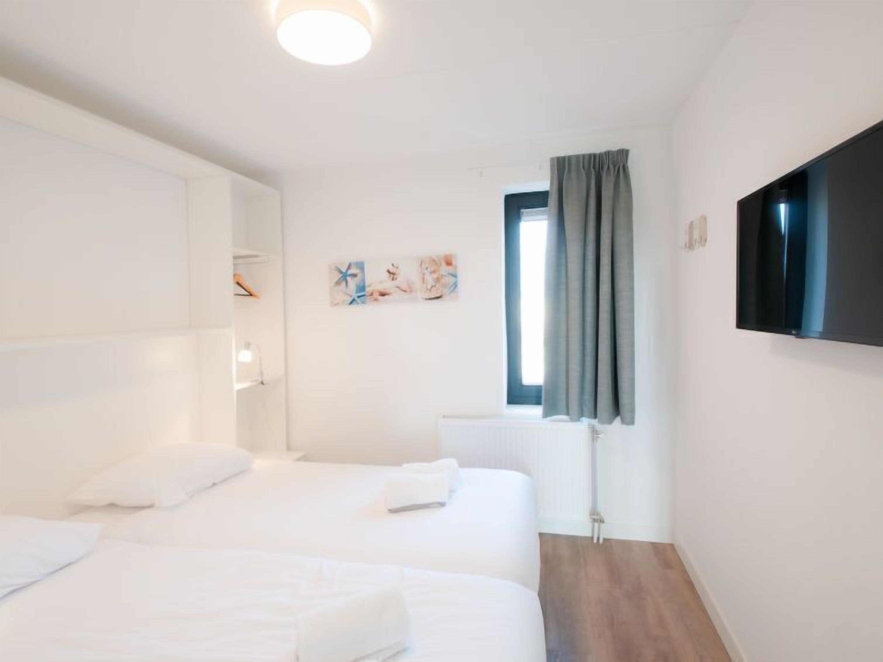 Ein Hotelzimmer mit zwei Betten, einem Flachbildfernseher, einem Fenster mit Vorhängen und Bildern an der Wand.