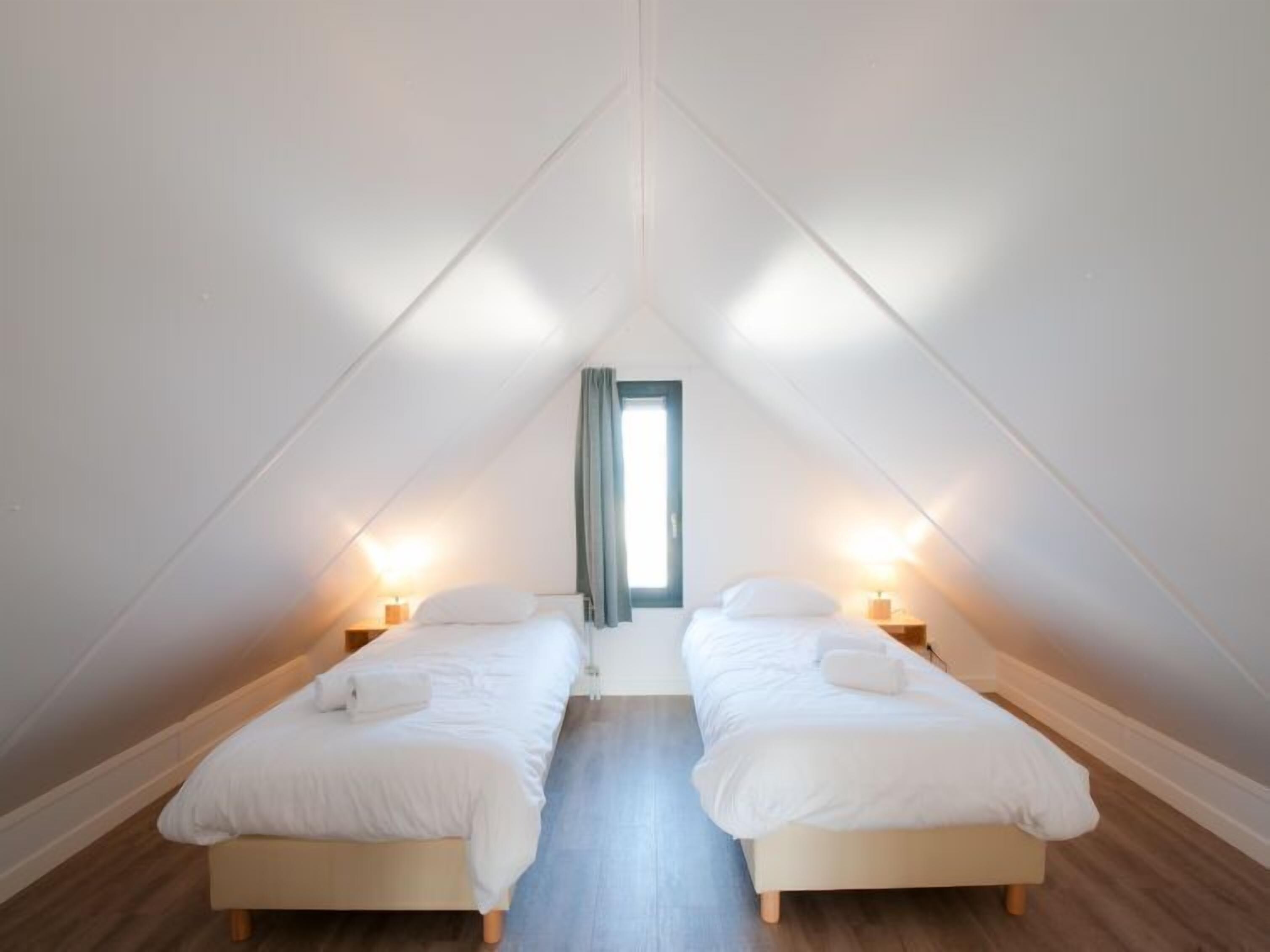 Une petite chambre mansardée, bien éclairée, avec deux lits simples, un plancher en bois et un puits de lumière.