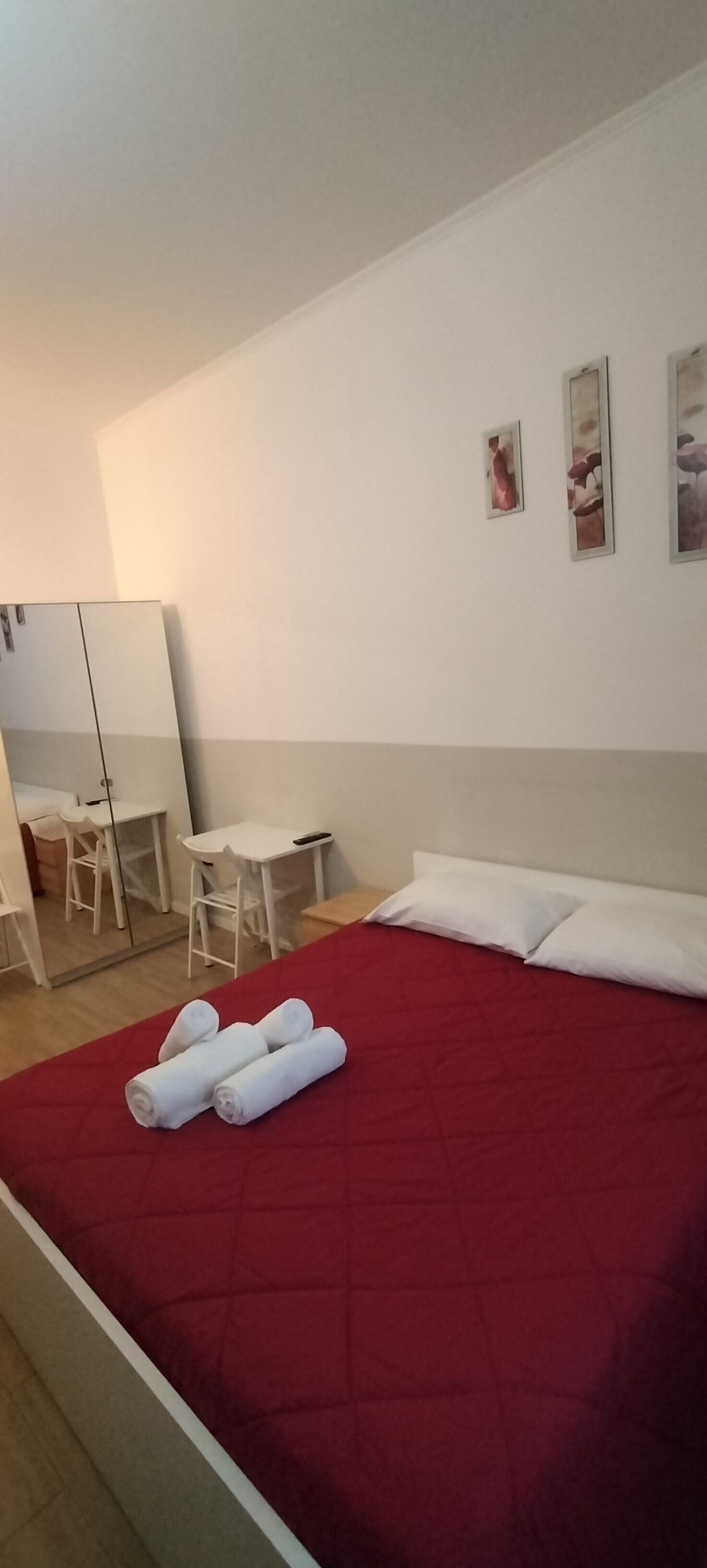 A hotel room with a red bedspread, a desk with a chair, a mirror, and three framed pictures on the wall.