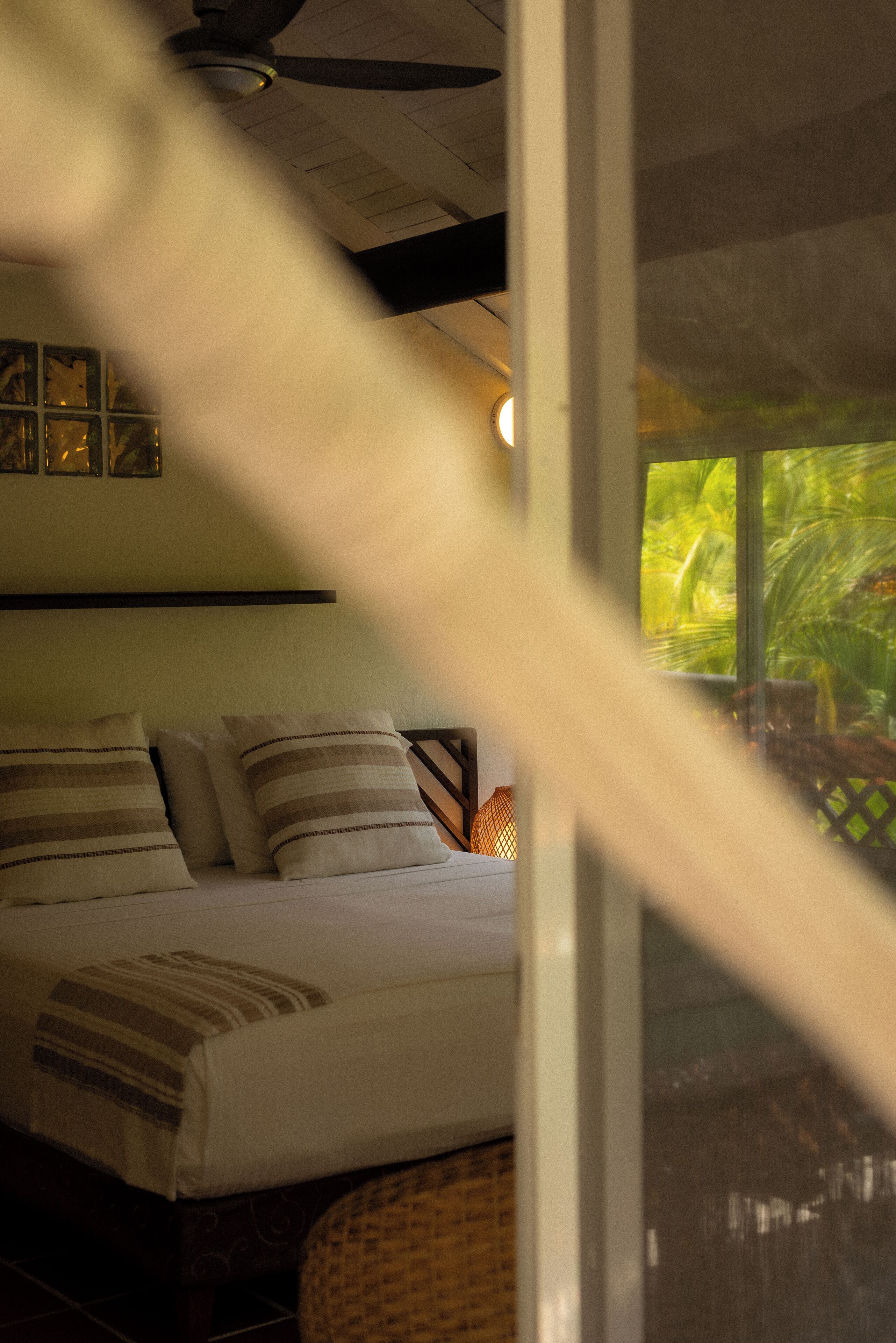 Ein Bett mit gestreiften Kissen, ein Deckenventilator und ein Fenster mit Blick auf Grünflächen.