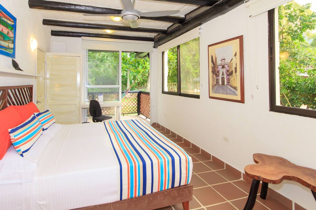 Un dormitorio con cama, almohadas, una silla, ventilador de techo y obras de arte en las paredes.