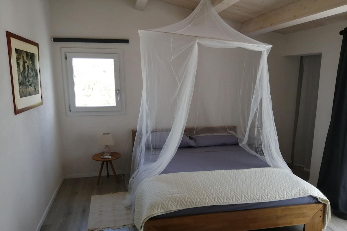 A bedroom with a wooden bed, white canopy, a small bedside table with a lamp, a window, and a framed picture on the wall.
