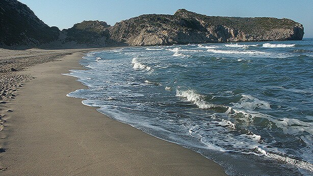 Una playa con un promontorio rocoso a lo lejos y olas rompiendo en la orilla.