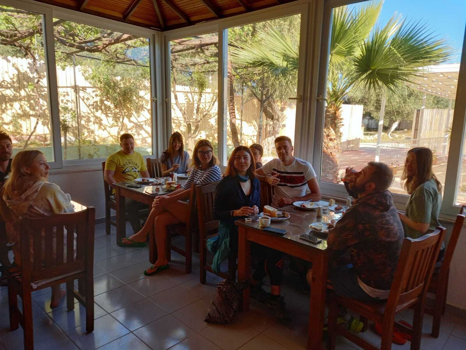 A group of people seated around a table in a room with large windows, wooden furniture, and a tiled floor.