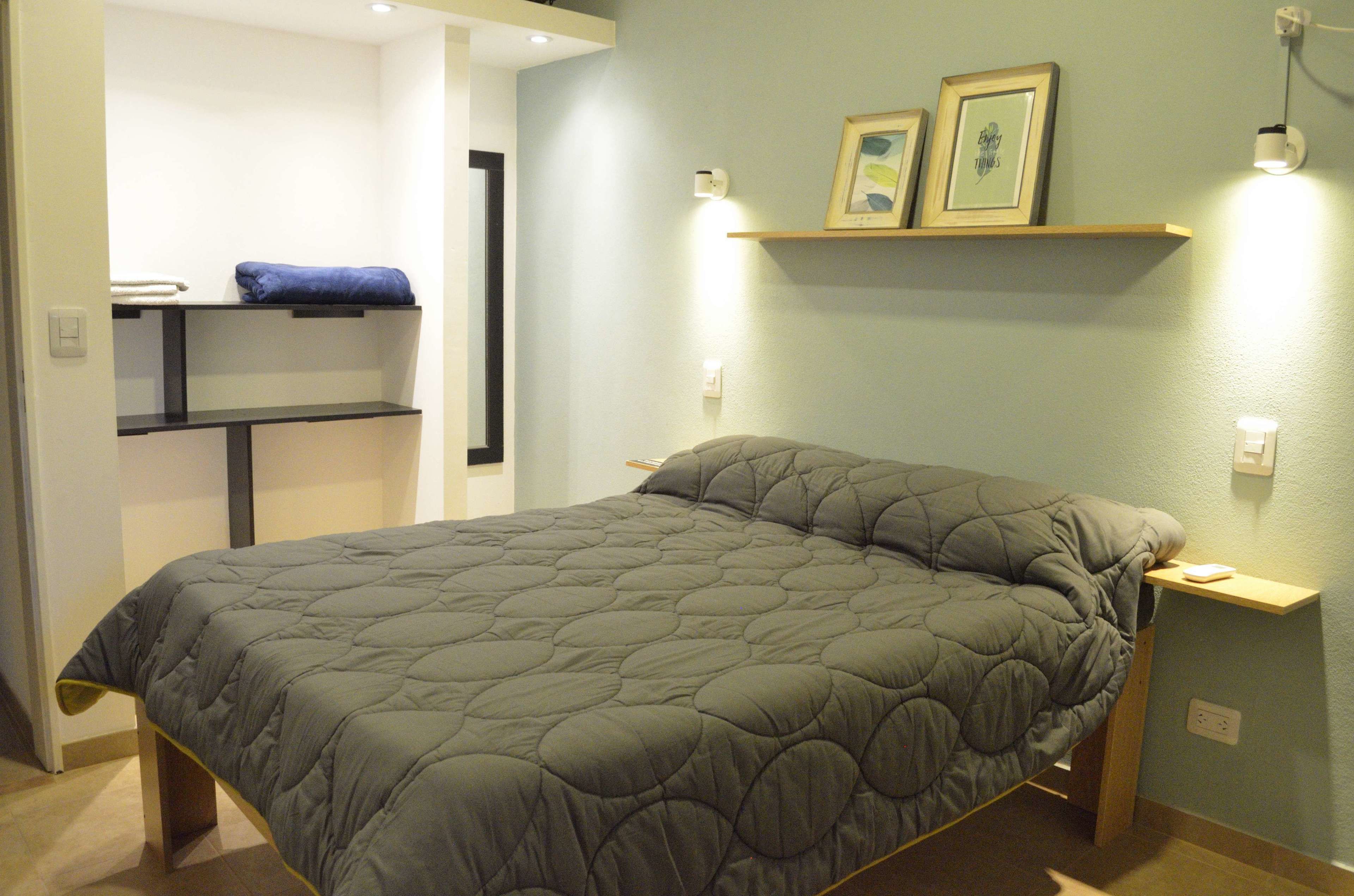 A neatly made bed with a grey quilt, a bedside table with a lamp, and a shelf with two framed pictures.