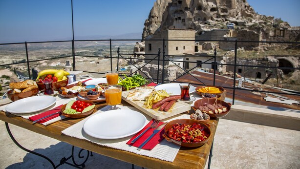 A dining table set with various foods and drinks, overlooking a rocky landscape with a large cave-like structure. - Ikarus Cappadocia Hotel (Nevsehir)
