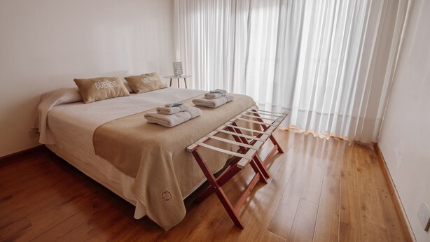 A neatly made bed with a wooden bench, a small table with a stack of books, and sheer curtains. - Casa Jainen (Ushuaia)