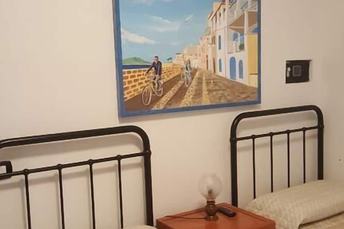 Una camera con due letti, un comodino con una lampada e un quadro raffigurante una costa.