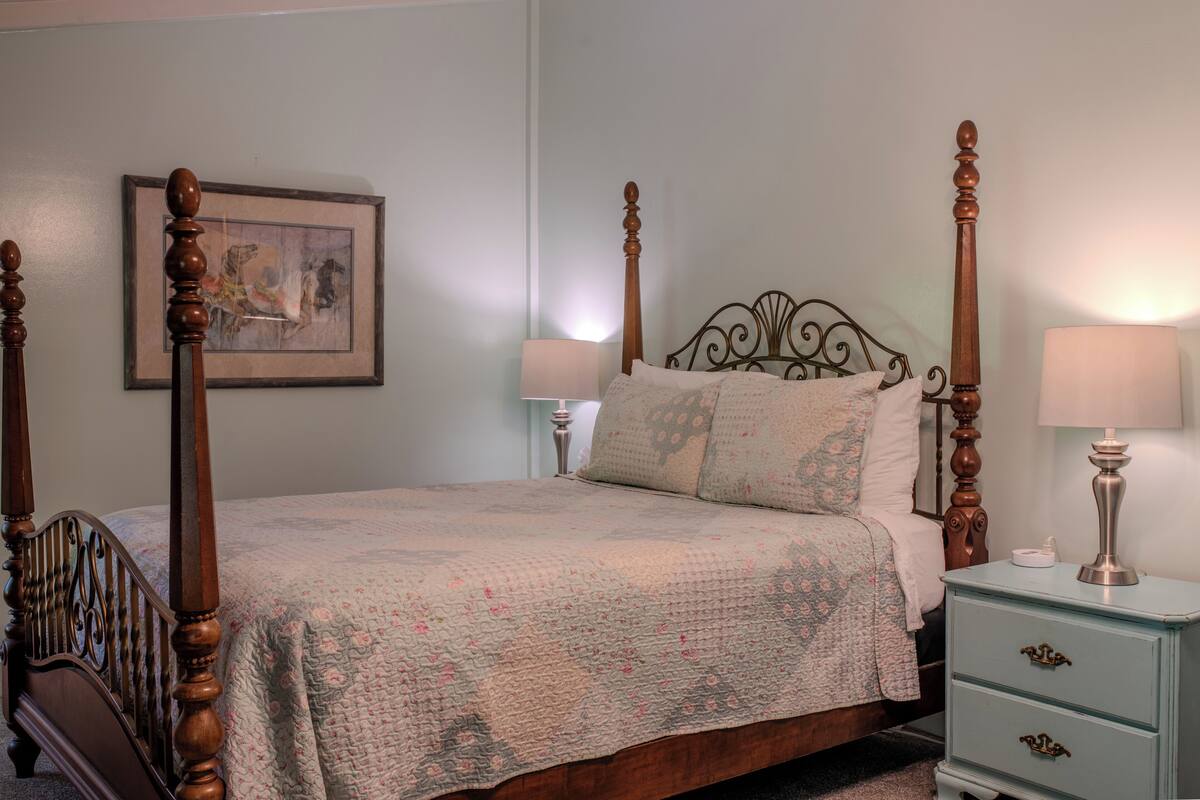 A four-poster bed with a quilt, a nightstand with a lamp, and a framed picture on the wall.
