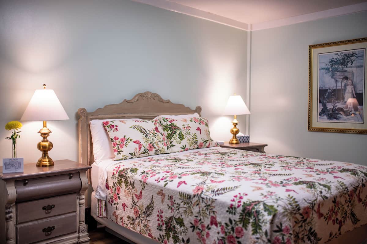 A bedroom with a floral bedspread, two bedside lamps, a nightstand with a vase of flowers, and a framed picture on the wall.