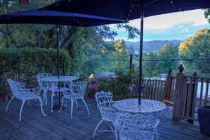 A deck with white metal chairs and tables, a dark umbrella, and a view of trees and mountains. - McKinley Edwards Inn (Bryson City)