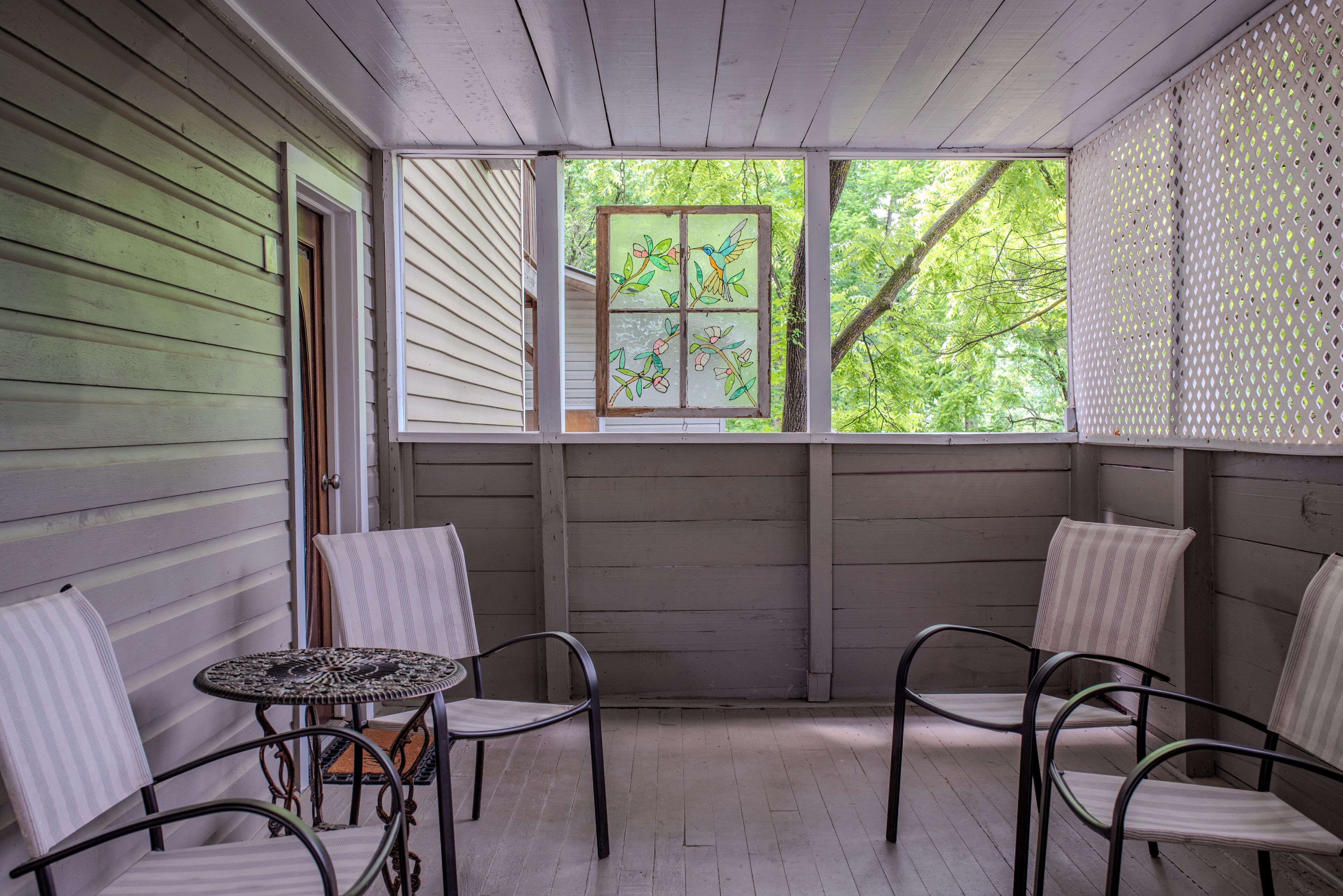 A covered porch with a table and chairs, a window with a stained glass design, and a view of trees.