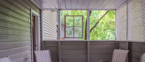 A covered porch with a table and chairs, a window with a stained glass design, and a view of trees.