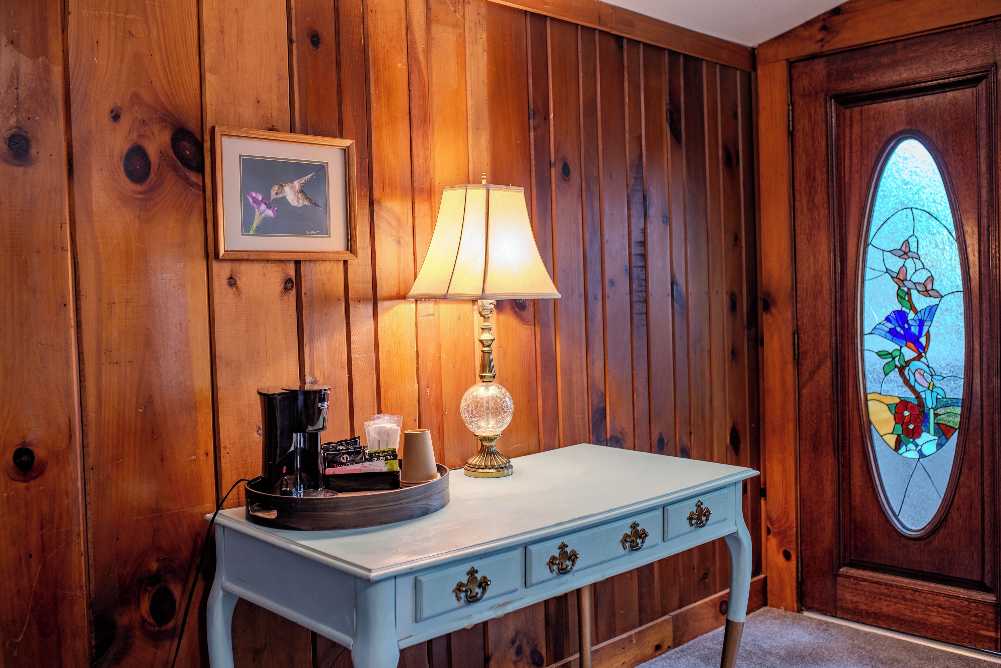 A wooden paneled wall, a white table with a lamp, a framed picture, and a stained glass window.