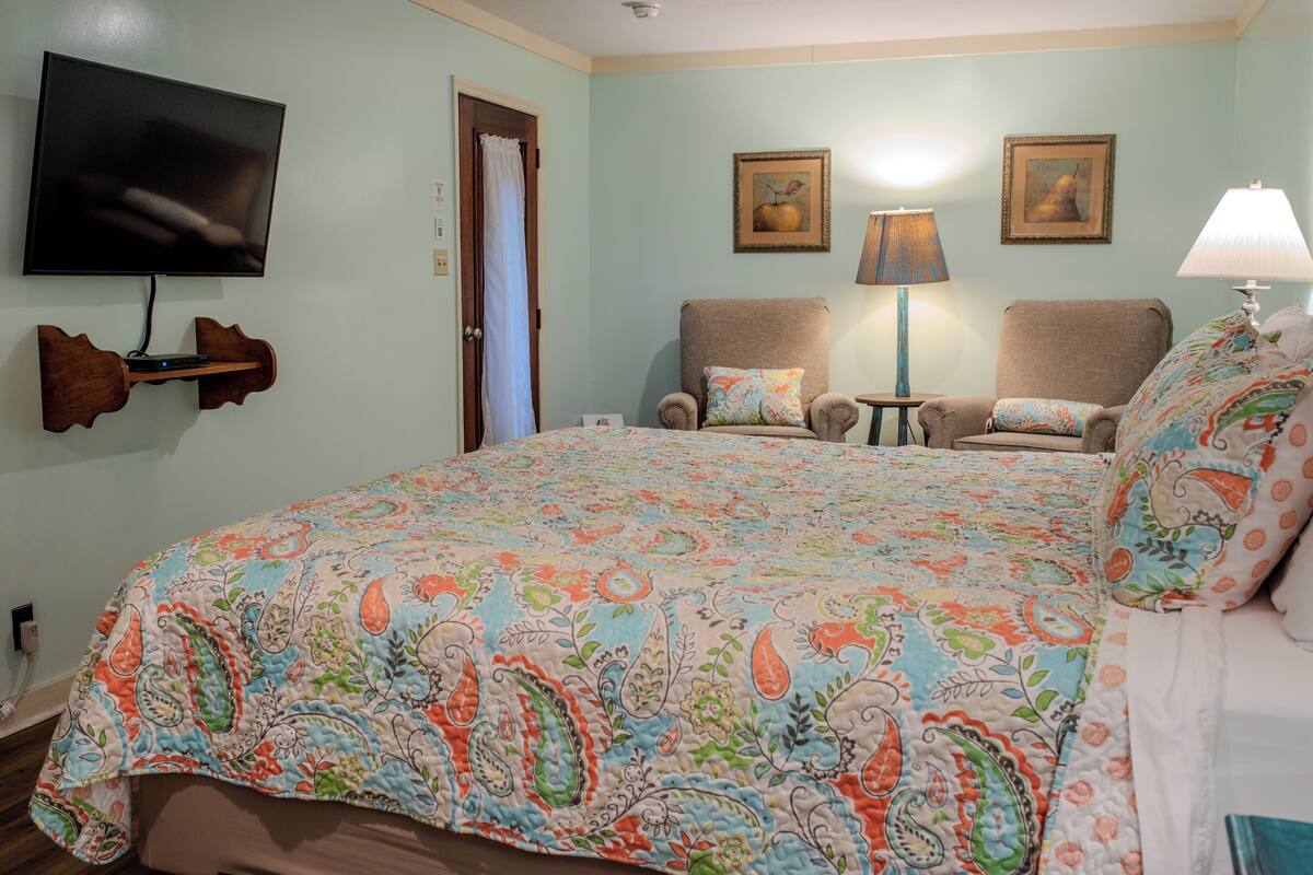A bedroom with a large bed, two armchairs, a TV mounted on the wall, and a door.