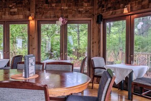 A wooden interior with a round table and chairs, large windows with a view of trees, and a hanging flower arrangement.