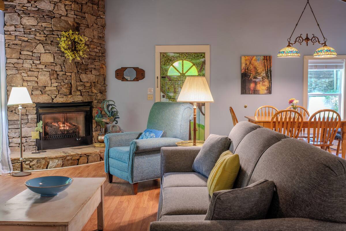 A living room with a stone wall, a fireplace, a blue armchair, a gray sofa, and a dining area with wooden chairs.
