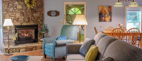 Una sala de estar con pared de piedra, chimenea, un sillón azul, un sofá gris y un comedor con sillas de madera.