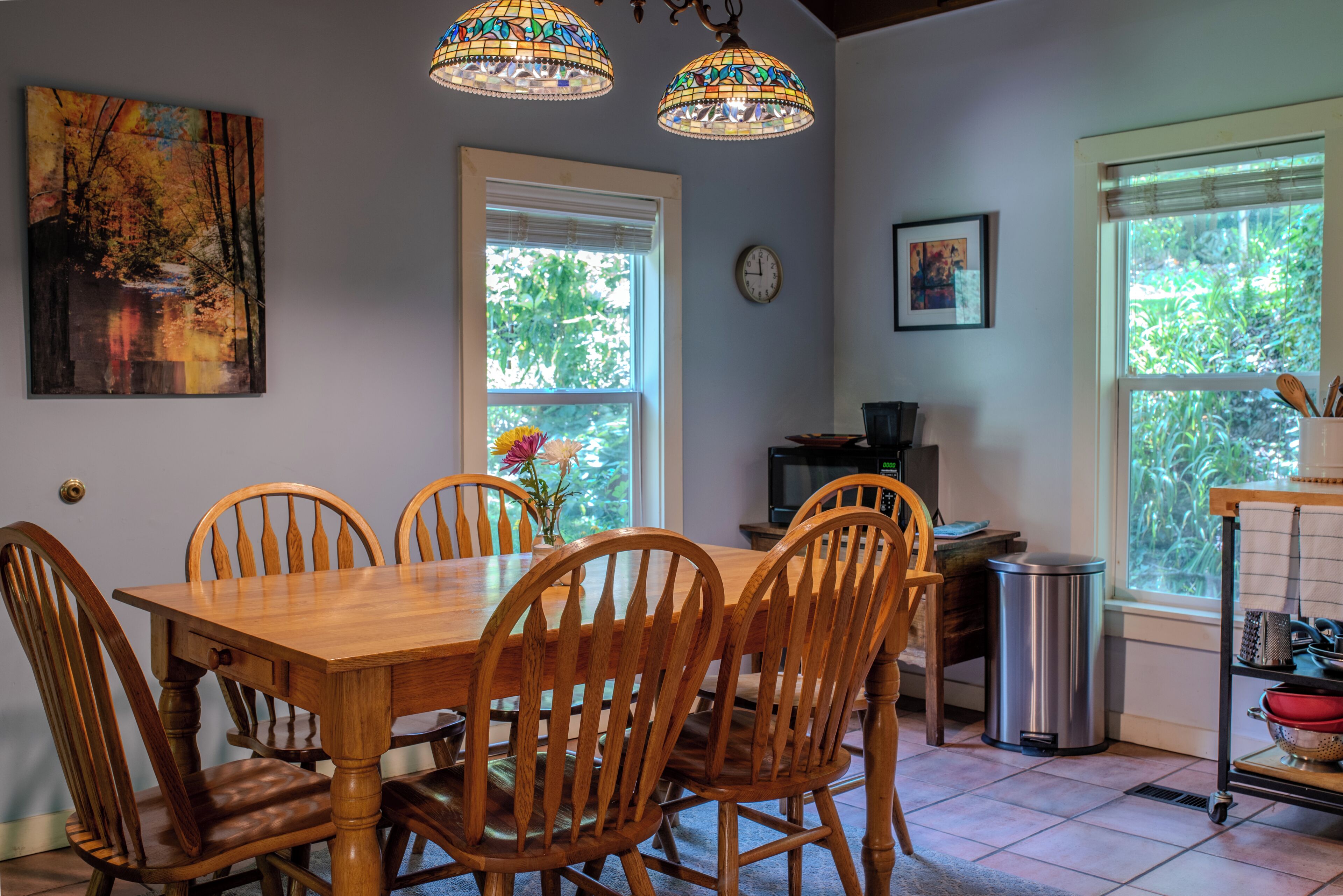 A dining area with a wooden table and chairs, a painting, a clock, and a window with a view of trees.
