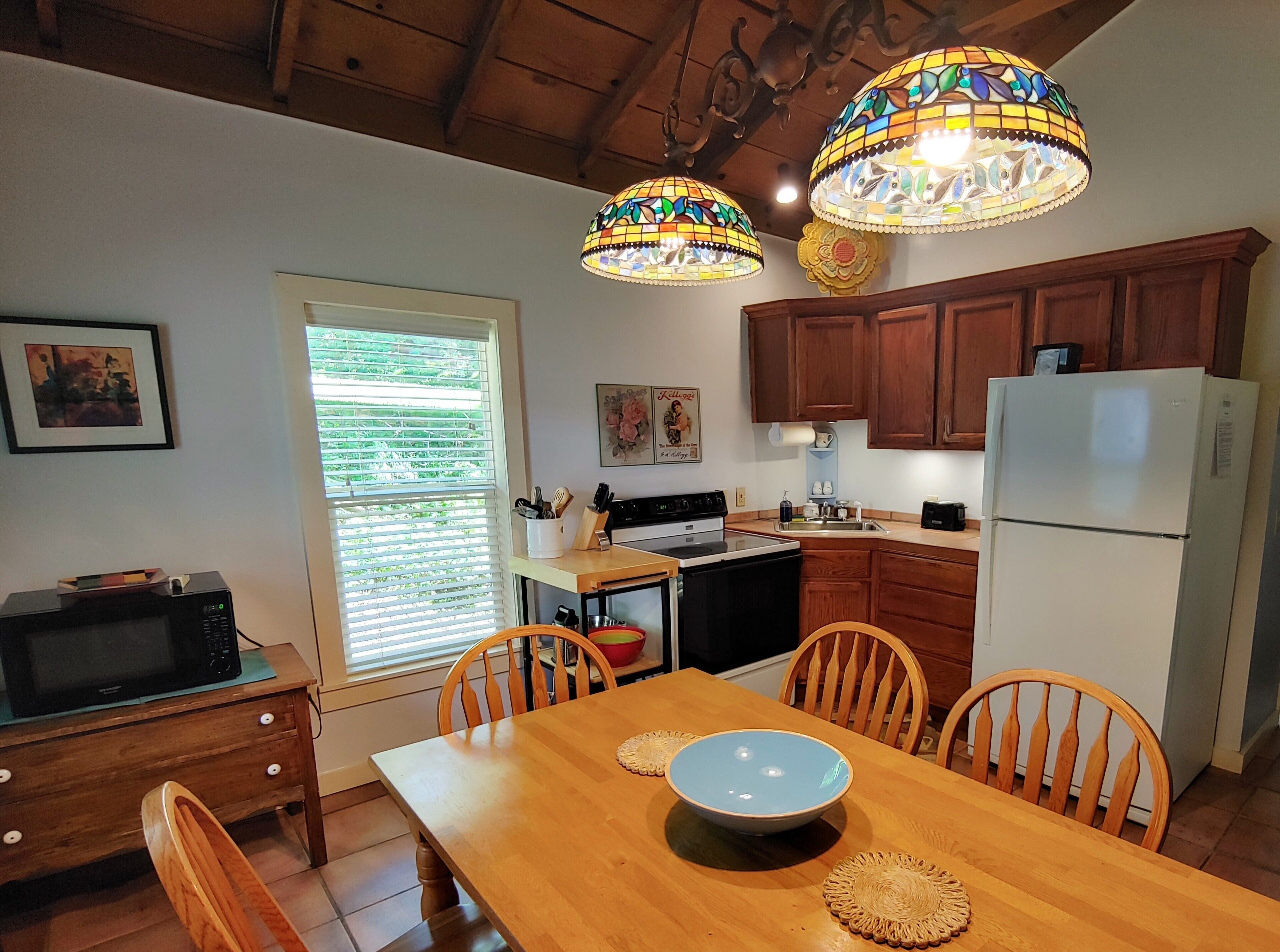 A kitchen with wooden cabinets, a refrigerator, a stove, and a dining table with chairs. There are two stained glass pendant lights above the table.