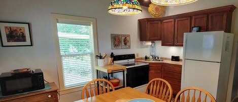 A kitchen with wooden cabinets, a refrigerator, a stove, and a dining table with chairs. There are two stained glass pendant lights above the table.
