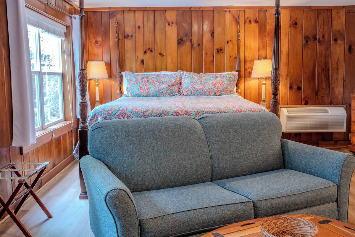 A bedroom with a bed, two bedside lamps, a wooden headboard, a window with curtains, and a wooden floor.