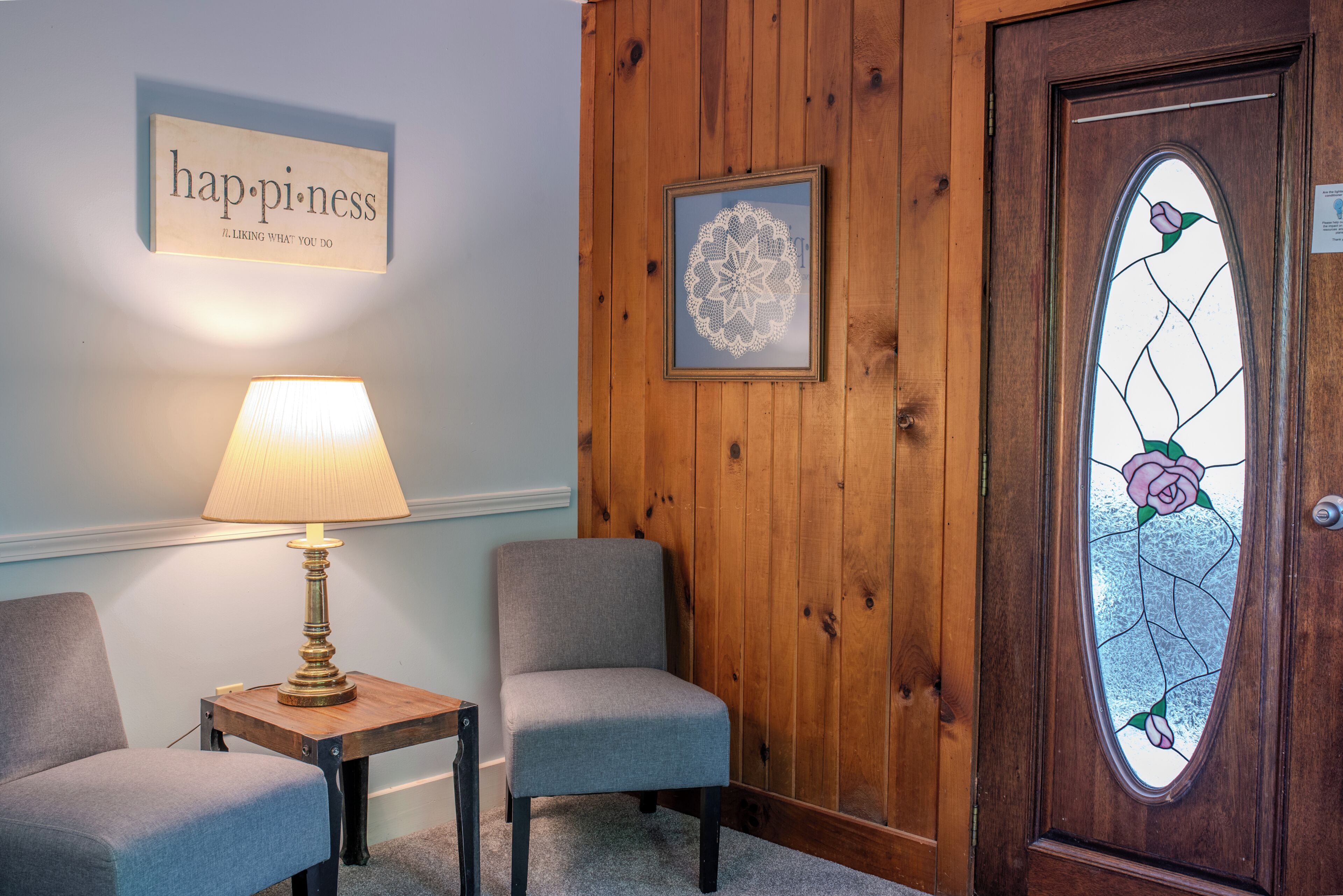 A room with a wooden paneled wall, a framed floral artwork, a lamp on a small table, and two chairs.