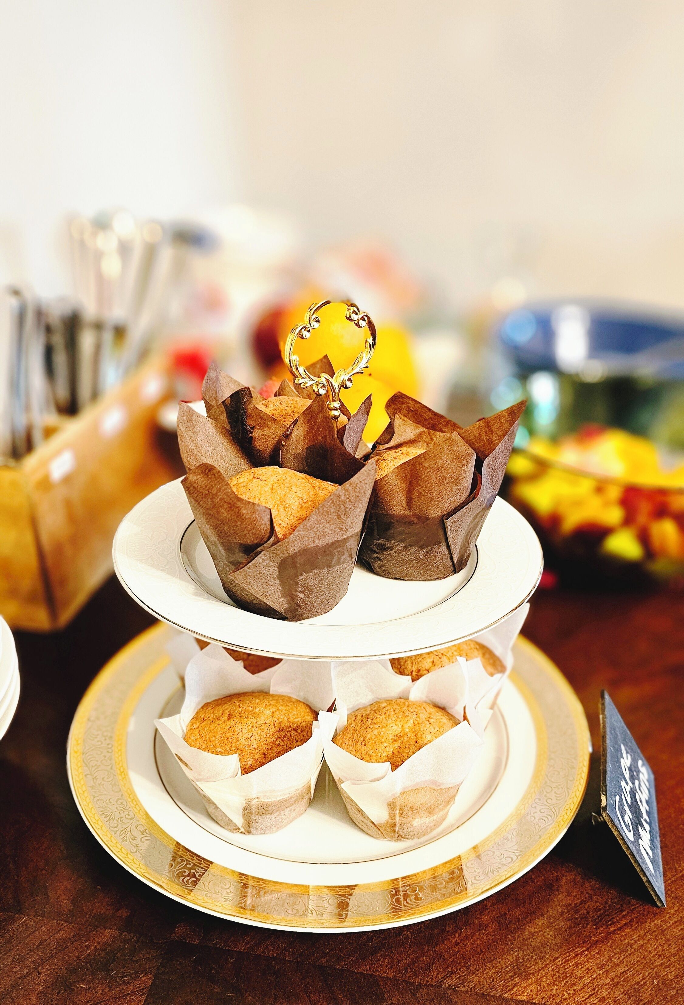 A tiered serving dish with pastries, a small sign, and a blurred background with a bowl of fruit.