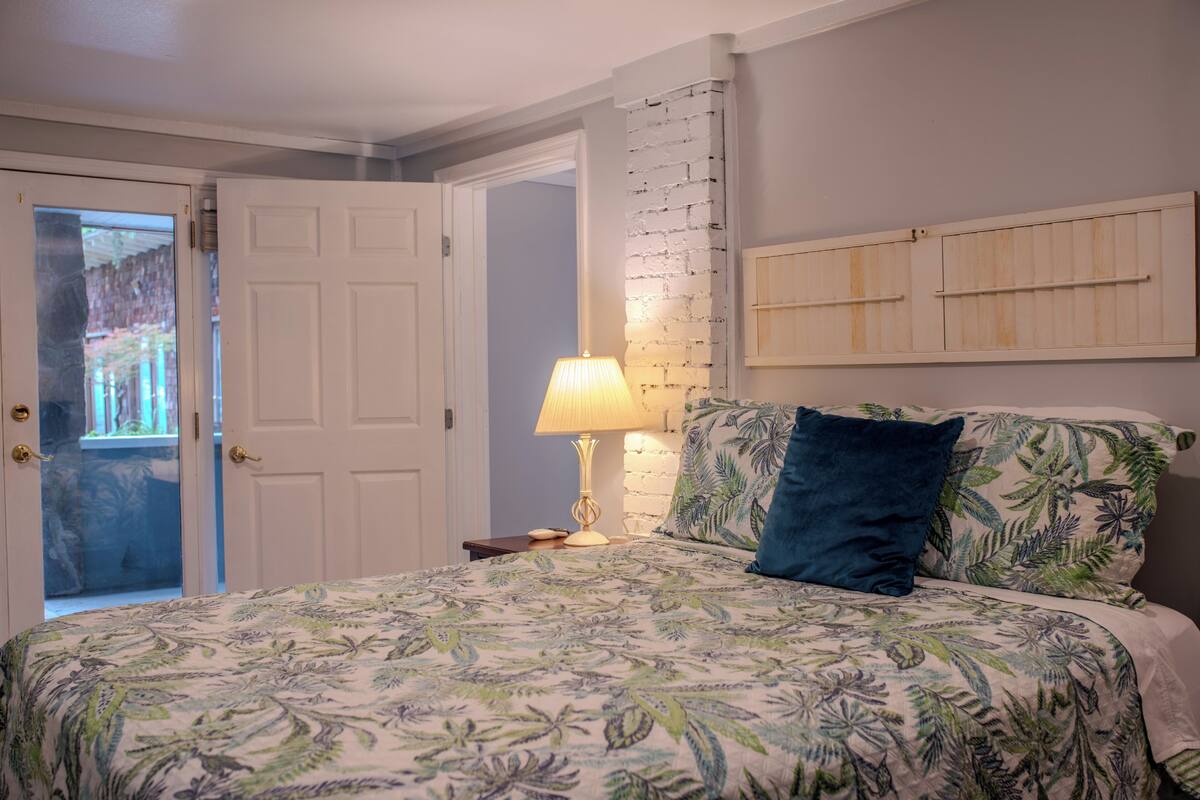 A bedroom with a bed, a nightstand, a lamp, a door, and a brick wall.