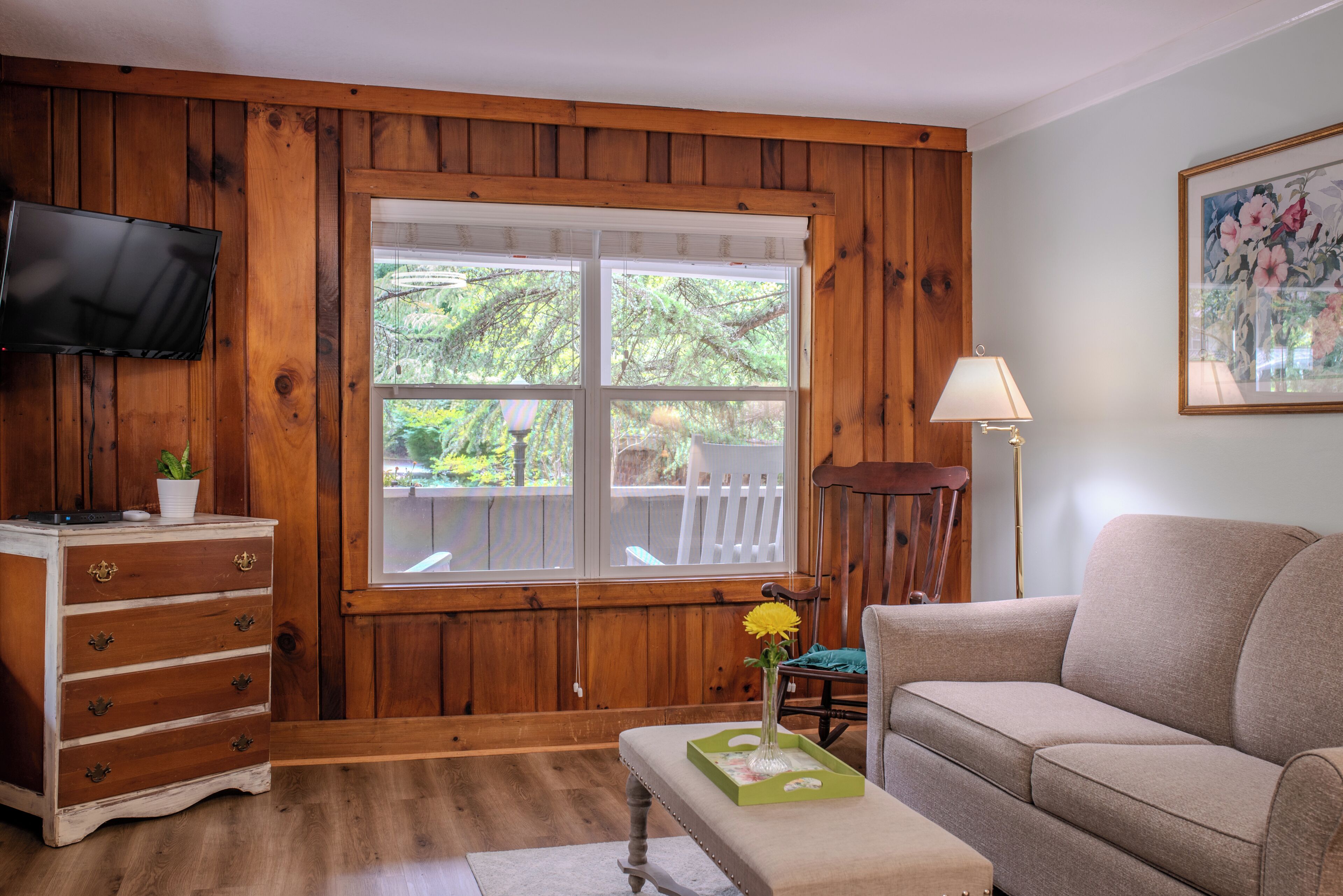 A room with wooden paneling, a television mounted on the wall, a window with a view of trees, a sofa, a chair, and a small table with a vase of flowers.