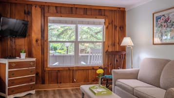A room with wooden paneling, a television mounted on the wall, a window with a view of trees, a sofa, a chair, and a small table with a vase of flowers.